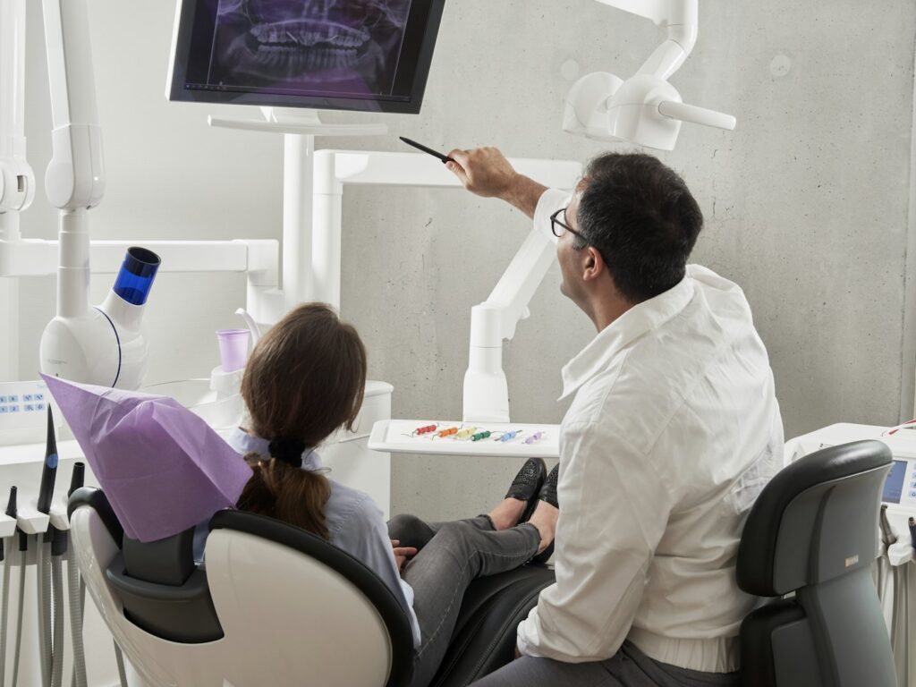 A dentist seated beside a patient, pointing at an X-ray displayed on a wall-mounted screen.